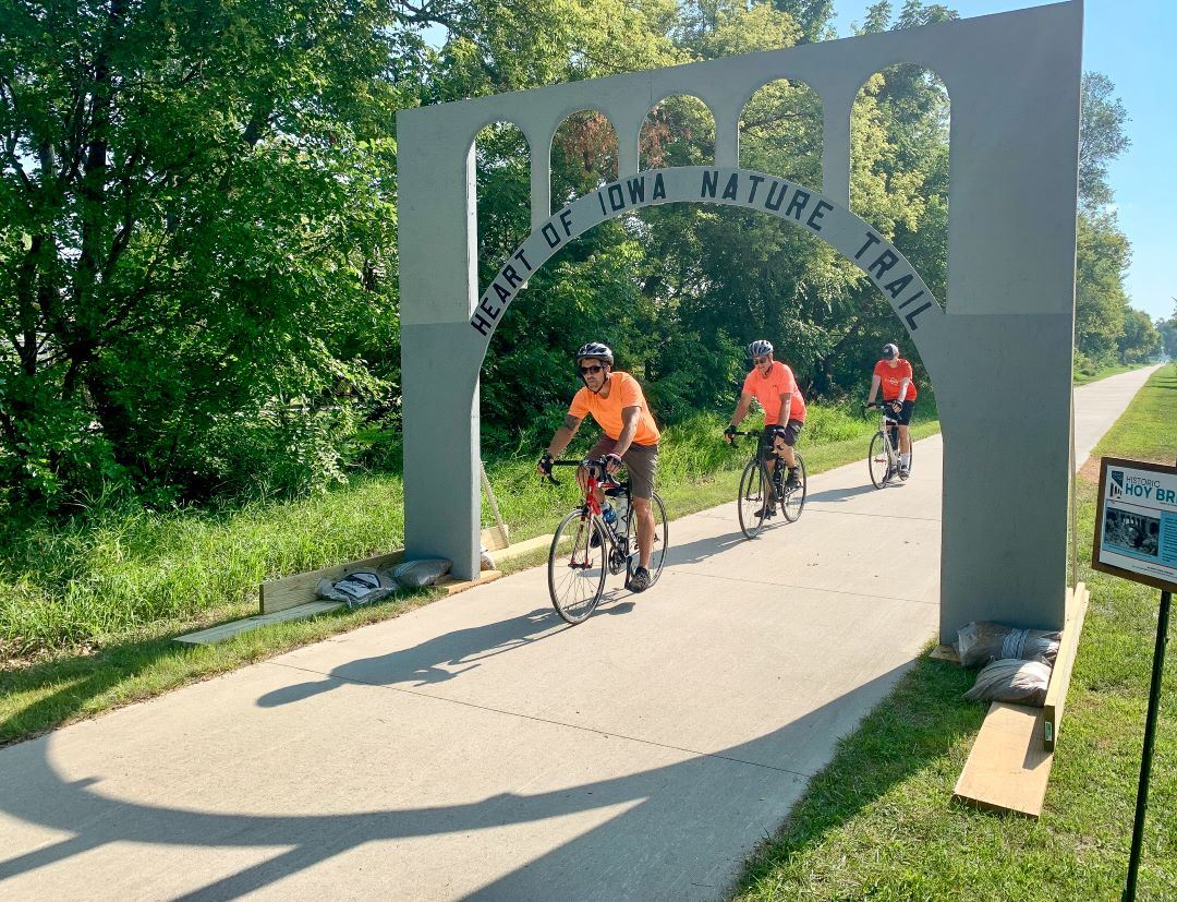 Cyclists wearing orange shirts ride under Hoy Bridge arch along paved Heart of Iowa Nature Trail