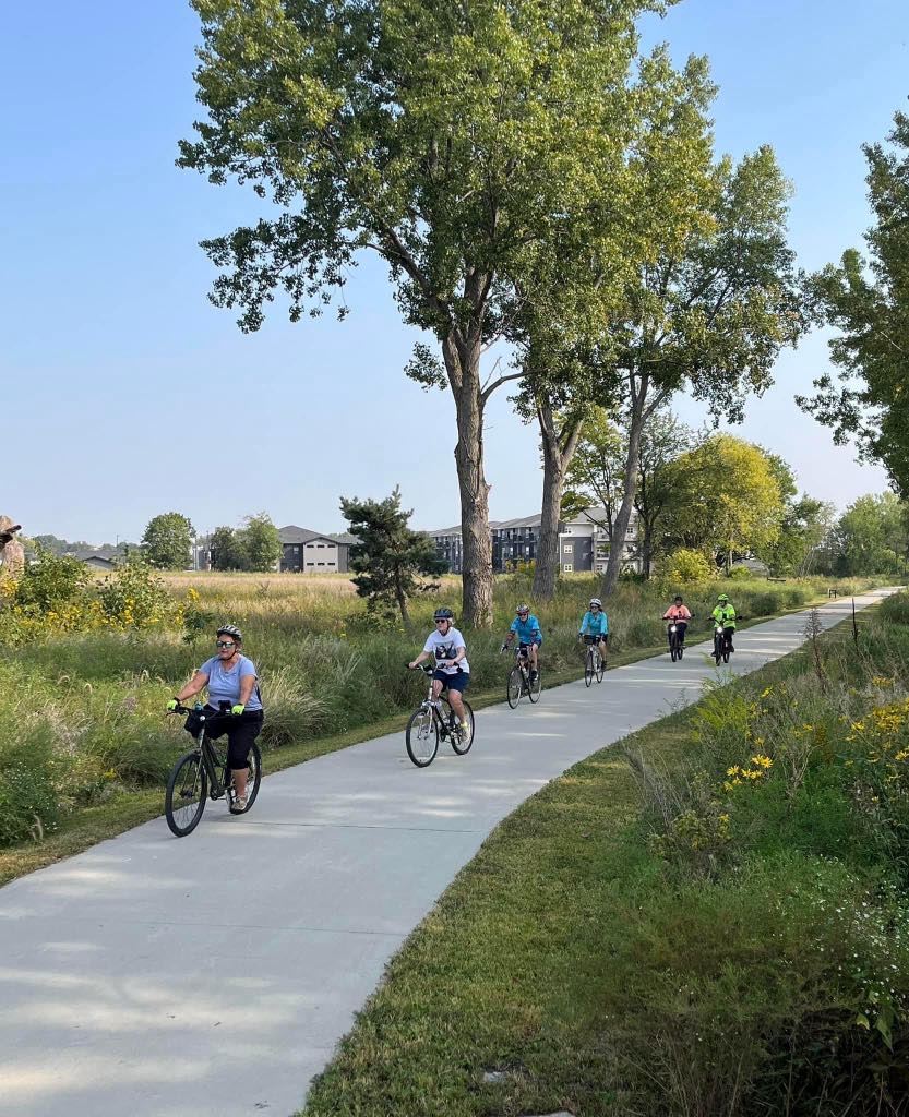 Line of six bicyclists ride toward viewer on paved trail with green grass and tall trees alongside