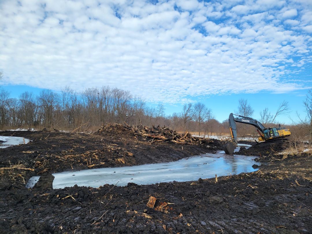 Excavator machine scoops into large depression in ground, filled with water, surrounded by mud